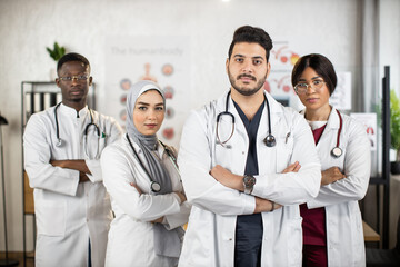 Team of multiracial doctors in white lab coats keeping hands crossed while standing together at...