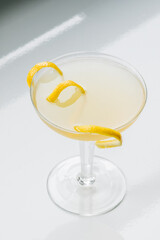 yellow cocktail in coupe glass with long curly lemon twist against white background
