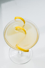 yellow cocktail in coupe glass with long curly lemon twist against white background