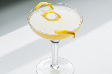 yellow cocktail in coupe glass with long curly lemon twist against white background