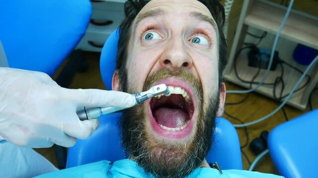 A young bearded man is afraid of professional brushing his teeth and screams in horror while sitting in the dentists chair