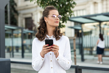 Smiling curly woman wearing trendy glasses walks down the central city street and uses her phone. Business woman using smartphone at commuter train station.