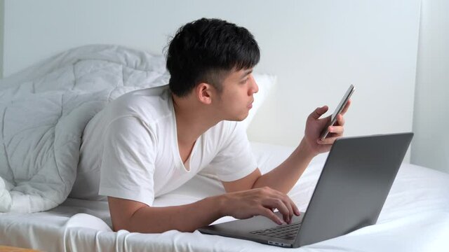 Young Asian man lying in the bed and using smartphone and laptop