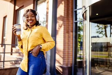 Portrait of young african american woman wearing yellow blouse smiling happy dancing listening to music using headphones drinking coffee at the city street outdoor.