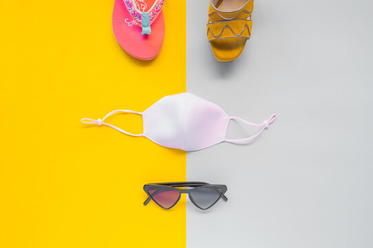Flip-flops, sunglasses, high-heeled sandals and a mask on a yellow and gray cardboard. Summer and coronavirus concept with different shoes neatly on a background divided in half. Top view, symmetry.