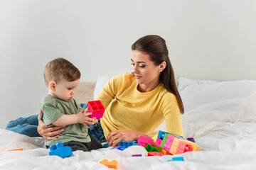 Obraz na płótnie Canvas Mother hugging toddler son with building blocks on bed