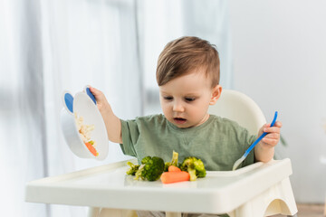 Toddler kid holding spoon and bowl near vegetables on high chair