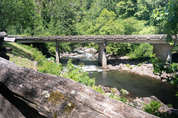 Fototapeta premium A rural elevated roadway bridge crossing a freshwater mountain stream ecosystem with moderately high white fast moving water flow after a summer thunderstorm as soaked the nearby hills 