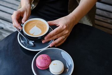 Fototapeta na wymiar Woman with a cup of aromatic coffee in her hands sits on the summer terrace of the restaurant. The girl drinks hot latte after breakfast with sweet macaroon dessert. Delicious French dessert macaroon