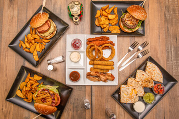 set of fast food dishes and hamburgers on a wooden table. Synchronized quesadillas, three forks, assorted starters, deluxe potatoes