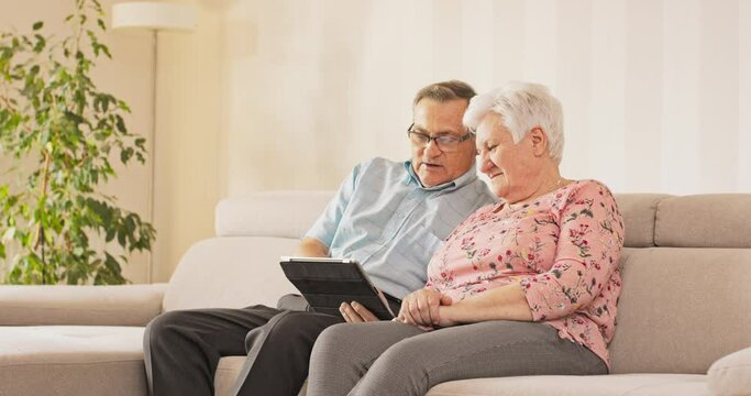 An aged couple sits on couch in living room, the husband sitting with glasses holds a tablet and taps his finger on it, the wife sitting next to him looks at the screen, they look at pictures, videos