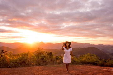 Young woman barefoot with arms raised and wearing a hat, enjoying nature, with the sunset in the background.