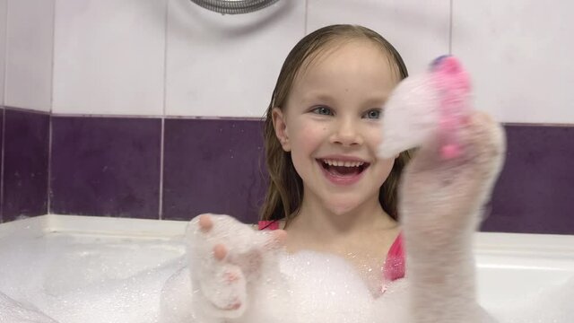 A little girl in a pink swimsuit blows the foam off her hands while playing with toys in the bath.