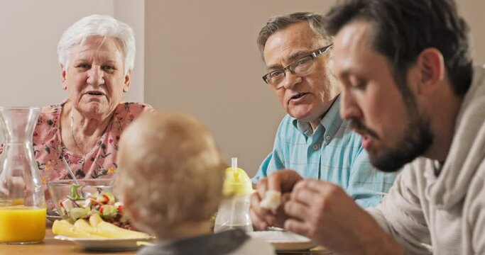 A brunette men sits at the table with his parents and feeds his little, cute, sweet son with blond hair, grandparents admire their grandson's appetite, they eat breakfast, family atmosphere