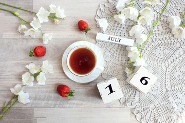  Calendar for July 16 : cubes with the number 16, the name of the month of July in English, a cup of tea on a gray openwork napkin, scattered flowers of bluebells and strawberries, top view