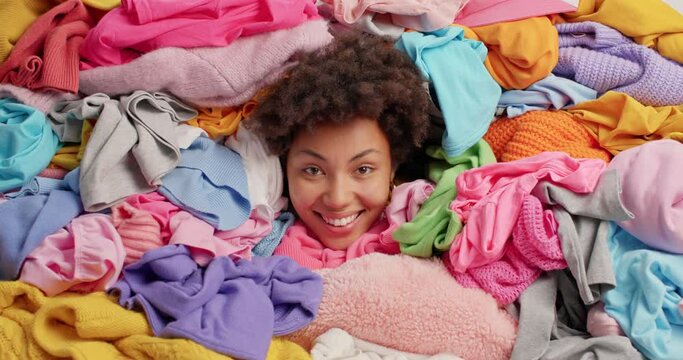 Positive African American woman stucks head in pile of multicolored laundry smiles gladfully surrounded by differet clothing cleans out closet collects items for donation. Housework concept.