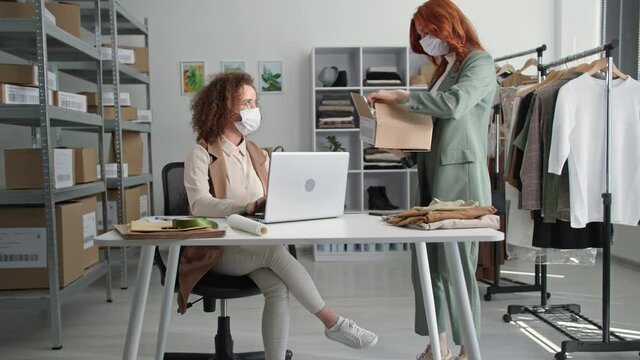 selling clothes online, young woman in medical mask together with female employee is shipping goods and packing bag in box sitting on warehouse in boutique