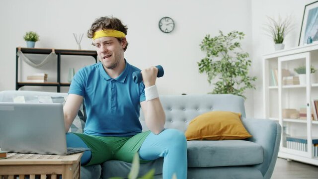Young man in colorful sportswear is exercising with dumbbell and using laptop enjoying online training at home. Sports and technology concept.