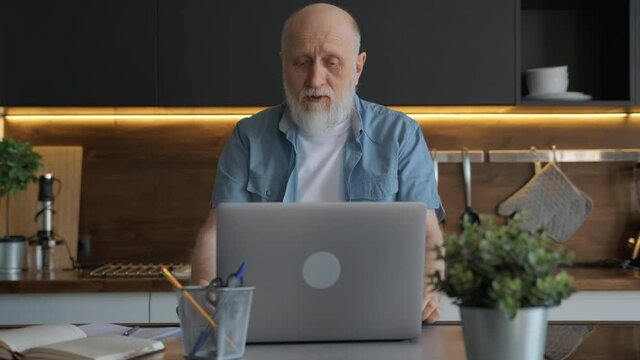 60-year-old elderly businessman works remotely from his home office talking on webcam sitting at table with laptop, solves financial issues with clients, takes online orders, develops online business.