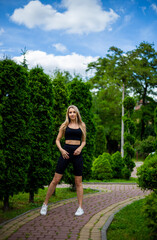 Obraz na płótnie Canvas Young beautiful sporty blonde woman in a black T-shirt and in black tight sports shorts in good shape posing in front of green trees
