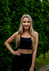 Naklejka na ściany i meble Young beautiful sporty blonde woman in a black T-shirt and in black tight sports shorts in good shape posing in front of green trees