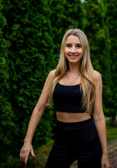 Young beautiful sporty blonde woman in a black T-shirt and in black tight sports shorts in good shape posing in front of green trees