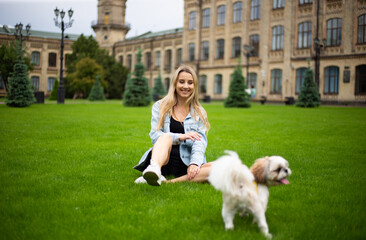 Young beautiful modern fashionable girl in a denim jacket sits on the grass and plays with a dog