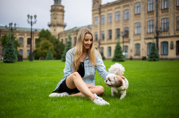 Young beautiful modern fashionable girl in a denim jacket sits on the grass and plays with a dog