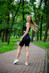 Young beautiful sporty blonde woman in a black T-shirt and in black tight sports shorts in good shape posing in front of green trees