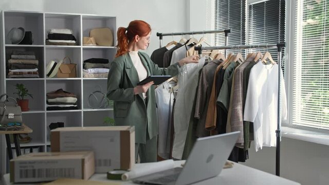 young business woman owner of clothing store with tablet in hands checks availability of goods on hangers, smiles and looks at camera