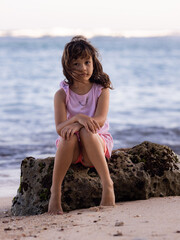 Young girl sitting on the rock near the ocean. Smiling girl. Happy childhood. Spending time on the beach. Vacation in Asia. Pandawa beach, Bali, Indonesia