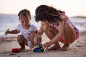 Baby boy and young girl sitting on sandy beach near the sea, playing with sand scoop and toy car....