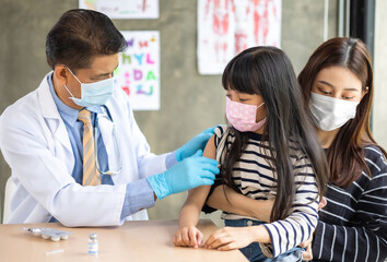 Asian  senior doctor wearing gloves and isolation mask is making a COVID-19 vaccination in the shoulder of child patient with her mother at hospital.