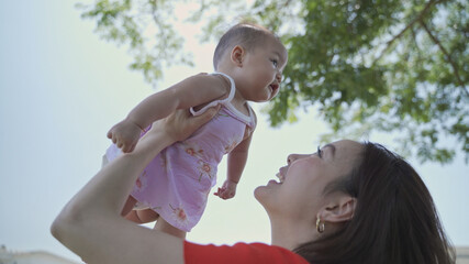 Obraz premium asian woman mother playing with happy baby infant in garden on sunny day under tree on picnic vacation. smile baby happy fun family time. raising in the air. baby flying in the sky air with parent.