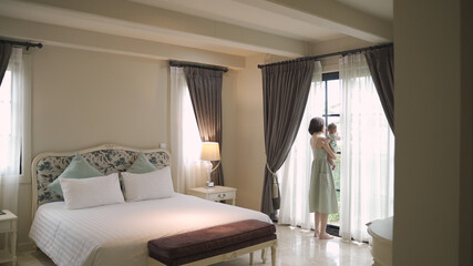 portrait of happy asian woman mother holding baby infant near window looking away at home. 4 months baby embracing in parent arm. relationship mother and daughter. mom carrying baby infant in bedroom
