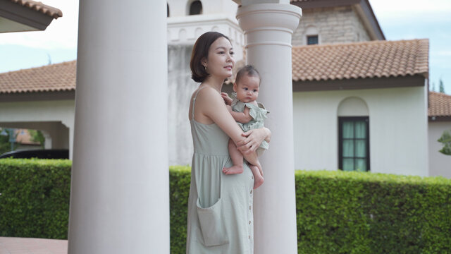 portrait of asian woman mother holding baby infant on balcony terrace at luxury home looking away the morning view. happy smile 30's female parent and 6 months baby at home near garden daytime.