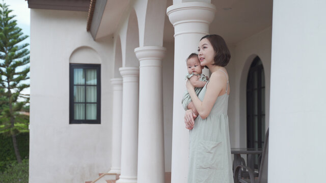portrait of asian woman mother holding baby infant on balcony terrace at luxury home looking away the morning view. happy smile 30's female parent and 6 months baby at home near garden daytime.
