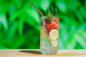 Summer cocktails, refreshing drinks concept. Drinking glass of hard seltzer cocktail with strawberry, mint lime and lemon outdoors. Infused water with fruit, green foliage on background.