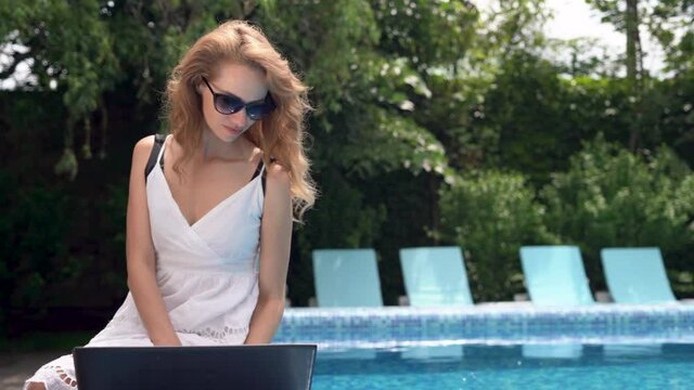 We work even on vacation Attractive young woman in sunglasses sits at the pool with a laptop. Cheerful smile.
