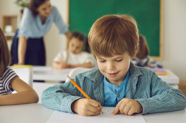 Enriching life with knowledge. Closeup portrait of little schoolboy elementary student writing or drawing in copy-book sitting at class desk with classmates and teacher on blurred background