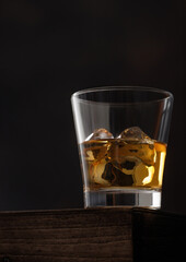 Glass of single malt whiskey with ice cubaes on top of wooden box on black