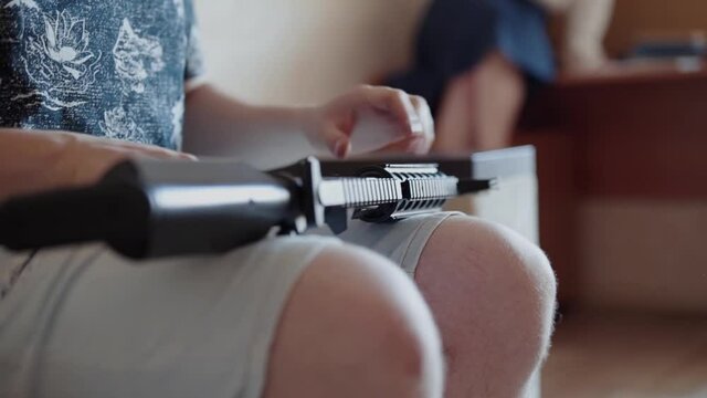 Caucasian man in shorts sits on the couch, holds black pneumatic assault rifle in his hands and examines it. Man puts machine gun on the sofa, close-up of the barrel.