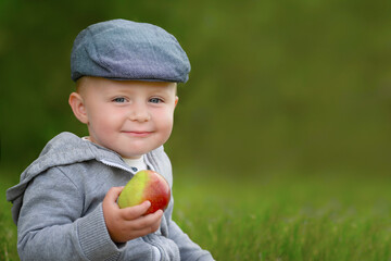 Boy in cap with apple in hand looking at camera. Blurred background. Copy space.