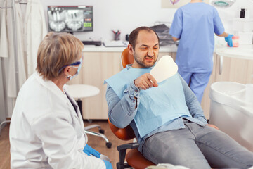 Obraz premium Sick patient inspecting teeth after dental surgery using mirror while sitting on stomatological chair in hospital clinic office. Senior stomatologist woman discussing about healdcare treatment