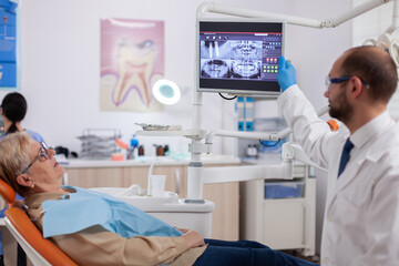 Naklejka na ściany i meble Stomatolog talking about teeh treatment to senior woman in dentist cabient. Medical teeth care taker pointing at patient radiography on screen sitting on chair.