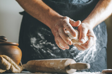Male hands knead dough with flour next to clay pot and oil bottle and rolling pin on dark table,...