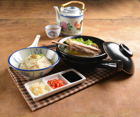 double boiled herbal bak kut teh pork ribs soup in black clay hot pot with rice and vermicelli noodle on wood table and sauces halal asian menu