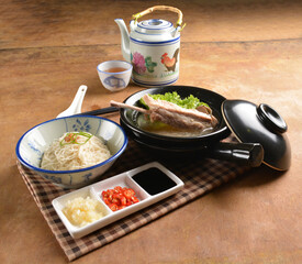 double boiled herbal bak kut teh pork ribs soup in black clay hot pot with rice and vermicelli noodle on wood table and sauces halal asian menu