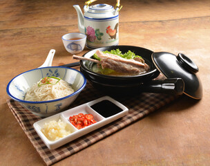 double boiled herbal bak kut teh pork ribs soup in black clay hot pot with rice and vermicelli noodle on wood table and sauces halal asian menu