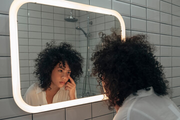 Young african american female checking her eye in the mirror. Putting on contact lenses in modern...